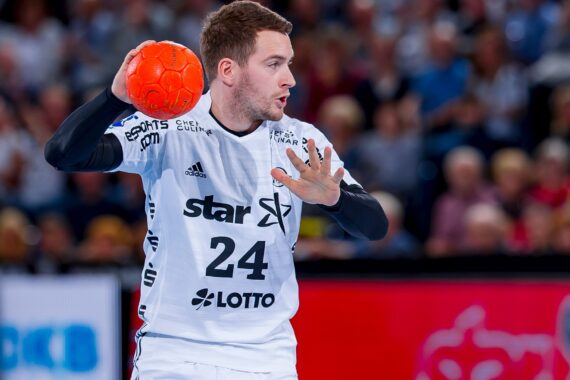 Ein männlicher Handballspieler in einem weißen Trikot mit der Nummer 24 bereitet sich darauf vor, einen orangefarbenen Ball während eines Spiels zu werfen. Im unscharfen Hintergrund sind Zuschauer zu sehen.