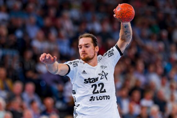 Ein männlicher Handballspieler in einem weißen Trikot springt in die Luft und bereitet sich darauf vor, einen orangefarbenen Ball zu werfen. Das Publikum im Hintergrund ist unscharf, was die intensive Aktion und die Konzentration des Spielers unterstreicht.