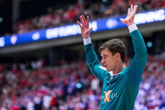 Ein männlicher Handballtorwart in einem blaugrünen Trikot steht mit erhobenen Armen und wirkt konzentriert. Der verschwommene Hintergrund zeigt eine überfüllte Arena mit rot gekleideten Zuschauern.