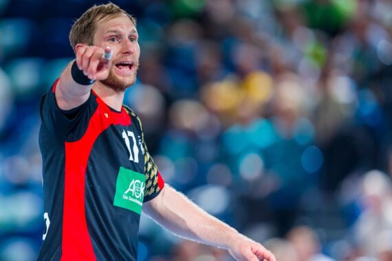 Ein männlicher Handballspieler in einem schwarz-roten Trikot mit der Nummer 17 zeigt nach vorne, den Mund geöffnet, als ob er schreien würde, während eines Spiels in einer Halle mit einem verschwommenen Publikum im Hintergrund.