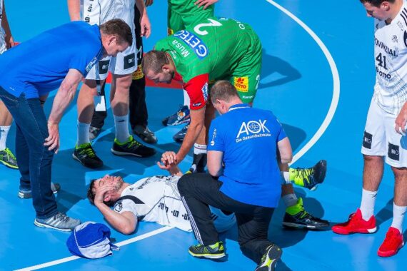 Eine Gruppe von Personen hilft einem verletzten Handballspieler, der auf dem Spielfeld liegt. Das medizinische Personal kümmert sich um ihn, während Mannschaftskameraden und ein anderer Spieler in der Nähe stehen und sich Sorgen machen.