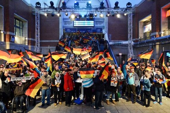 Eine große Menschenmenge versammelt sich drinnen, viele schwenken deutsche Fahnen und tragen deutsche Farben und feiern enthusiastisch. Eine Rolltreppe mit noch mehr Menschen führt zu einem hell erleuchteten Bühnenbereich im Hintergrund.