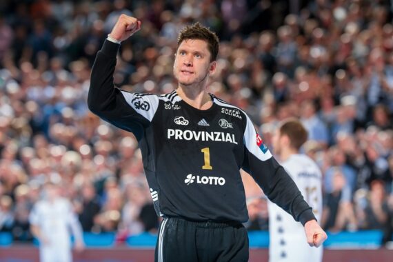 Ein männlicher Handballspieler in einem schwarzen Trikot mit der Nummer 1, der auf einem Spielfeld jubelnd die Faust hebt, mit einer verschwommenen Menschenmenge im Hintergrund.
