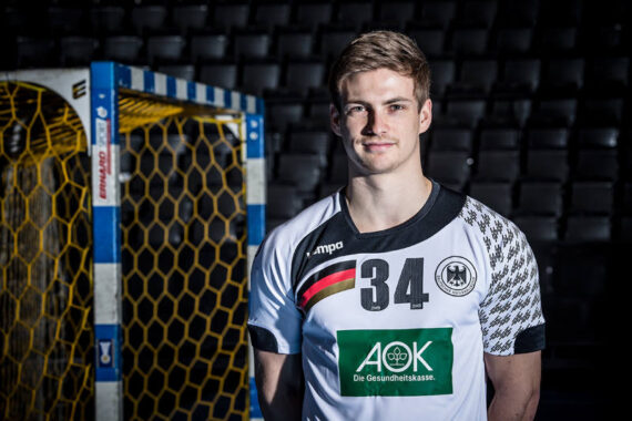 Ein männlicher Handballspieler in einem weiß-schwarzen Trikot mit der Nummer 34 steht vor einem Tornetz in einer Halle mit dunklen, leeren Sitzen im Hintergrund.