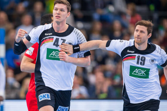 Zwei männliche Handballspieler in weißen und schwarzen Deutschland-Trikots feiern auf dem Spielfeld. Der Spieler mit der Nummer 47 auf der linken Seite hebt die Faust, während der Spieler mit der Nummer 19 auf der rechten Seite eine Hand auf den Arm seines Mitspielers legt.