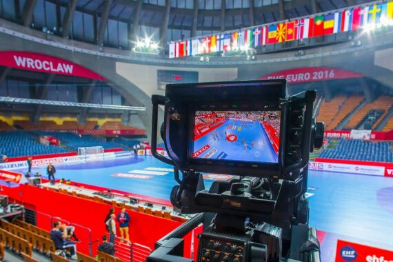Eine Kamera filmt eine hell erleuchtete Handballhalle mit blauem Bodenbelag, leeren Sitzen und verschiedenen internationalen Flaggen, die darüber hängen. Das Kameradisplay zeigt eine Nahaufnahme des Handballfeldes.