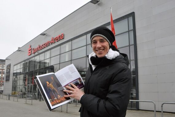 Eine Person in Wintermantel und Mütze hält an einem bewölkten Tag lächelnd eine Zeitschrift vor der Sparkassen-Arena auf.