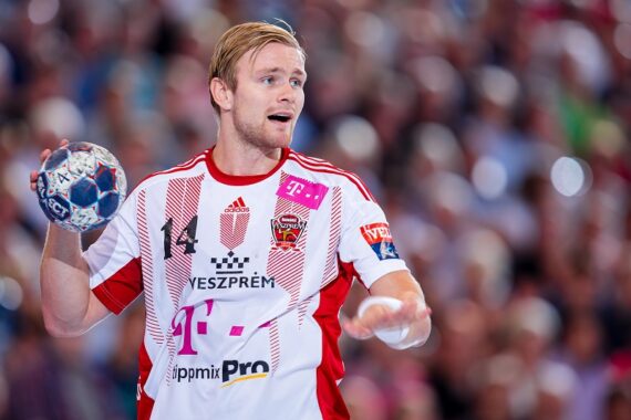 Ein männlicher Handballspieler in einem weiß-roten Trikot hält während eines Spiels einen Handball in der rechten Hand, mit einer unscharfen Menschenmenge im Hintergrund.