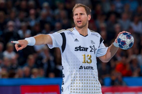 Ein männlicher Handballspieler in einem weißen Trikot mit der Nummer 13 hält während eines Spiels einen Ball in der einen Hand und zeigt mit der anderen auf den Ball, während im Hintergrund eine unscharfe Menschenmenge zu sehen ist.