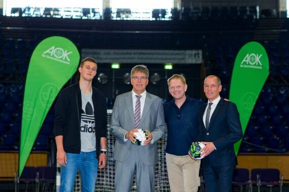 Vier Männer stehen in einer Halle auf einem Sportplatz, zwei halten Fußbälle. Im Hintergrund sind zwei grüne Werbebanner mit dem AOK-Logo und leere blaue Stadionsitze zu sehen.