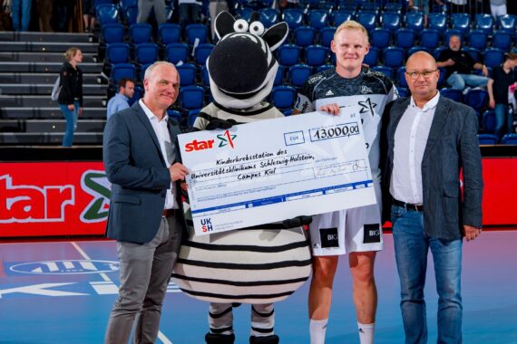 Vier Personen, darunter ein Zebramaskottchen, halten auf einem Sportplatz einen großen Spendenscheck über 43.000 Euro, während zwei Männer in Anzügen und ein Mann in Sportkleidung in die Kamera lächeln. Im Hintergrund sind blaue Stadionsitze zu sehen.