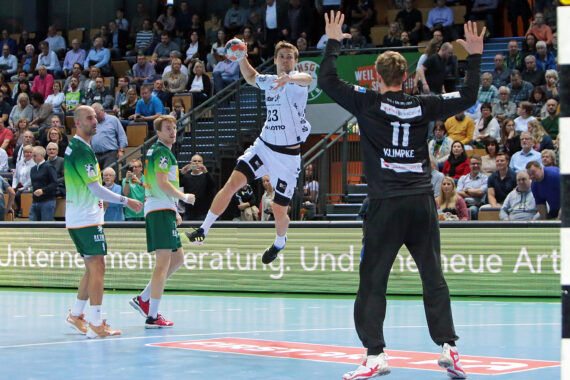 Ein Handballspieler in einem weißen Trikot springt zum Schuss, während ein Torwart in Schwarz die Arme zum Abwehren hebt. Zwei gegnerische Spieler in Grün und Weiß schauen zu. Das Geschehen spielt sich in einer überfüllten Sporthalle ab.