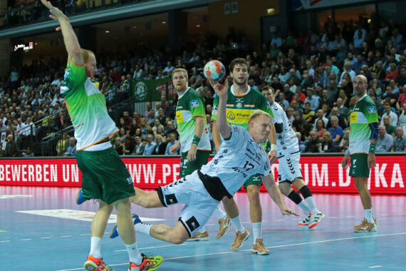 Ein Handballspieler in einem weißen Trikot taucht ab, um den Ball in Richtung Tor zu schießen, während ein Verteidiger in Grün den Arm zum Abwehren hebt. Im Hintergrund sind andere Spieler und eine volle Zuschauermenge zu sehen.