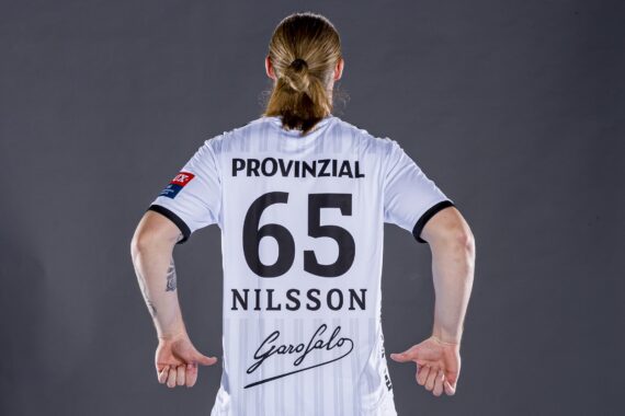 Eine Person mit zurückgebundenen langen Haaren, von hinten gesehen, trägt ein weißes Sporttrikot mit dem Namen NILSSON, der Nummer 65 und mehreren Logos, darunter PROVINZIAL, auf einem schlichten dunklen Hintergrund.
