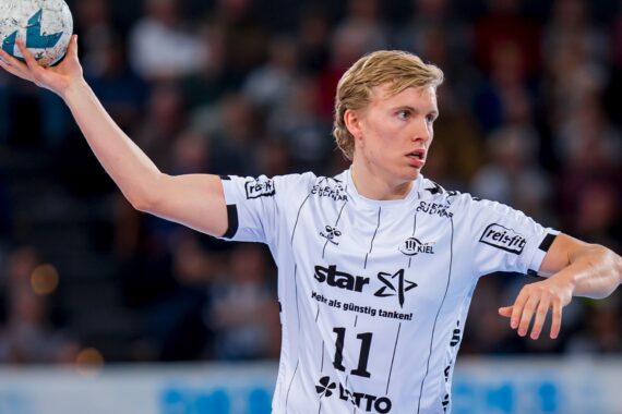 Ein männlicher Handballspieler in einem weißen Trikot mit der Nummer 11 bereitet sich darauf vor, den Ball während eines Spiels zu werfen, mit einer unscharfen Menschenmenge im Hintergrund.