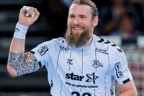Ein bärtiger männlicher Handballspieler in einem weißen Trikot mit schwarzen Mustern lächelt und hebt jubelnd die Faust. Er hat einen tätowierten Ärmel auf seinem linken Arm und steht auf einem Hallenplatz.