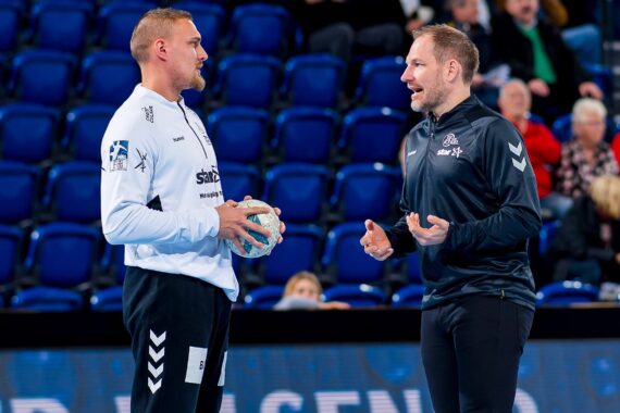 Zwei Männer in Sportkleidung stehen auf einem Hallenplatz und diskutieren. Der eine hält einen Handball, während der andere mit den Händen gestikuliert. Im Hintergrund sind blaue Stadionsitze und Zuschauer zu sehen.