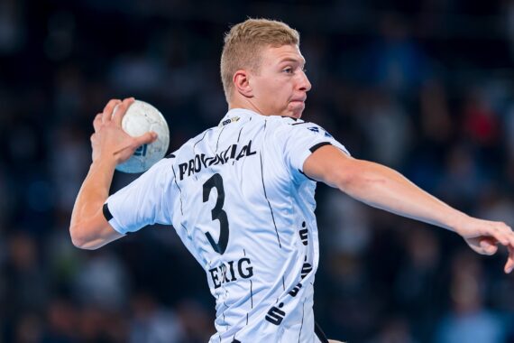 Ein männlicher Handballspieler in einem weißen Trikot mit der Nummer 3 bereitet sich darauf vor, den Ball während eines Spiels zu werfen. Der Hintergrund ist unscharf und zeigt eine Halle mit Zuschauern.
