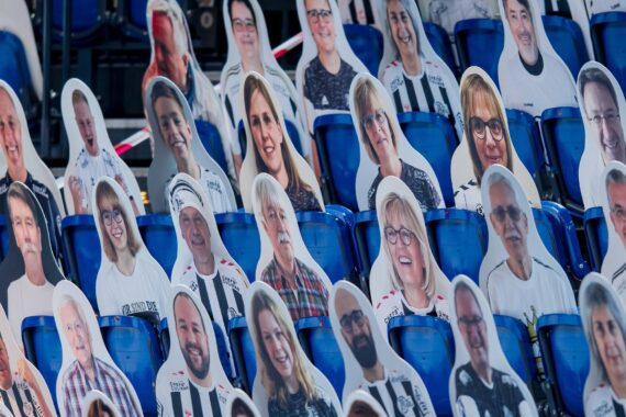 Reihen von blauen Stadionsitzen, gefüllt mit Pappfiguren von Menschen, jede mit einem anderen lächelnden Gesicht, die den Eindruck einer Menschenmenge bei einer Veranstaltung erwecken.