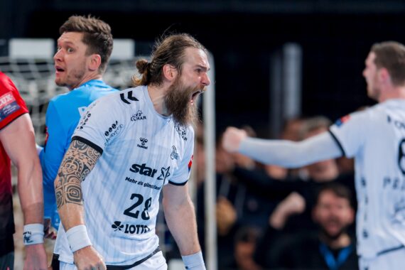 Ein Handballspieler mit langem Bart und Tätowierungen, der ein weißes Trikot trägt und leidenschaftlich schreit, steht vor einem anderen Spieler in Blau in der Nähe eines Tors, mit einer verschwommenen jubelnden Menge im Hintergrund.