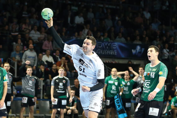 Ein Handballspieler in einem weißen Trikot springt und wirft den Ball während eines Spiels, umgeben von Spielern in grünen Trikots, während die Zuschauer im Hintergrund von der Tribüne aus zusehen.