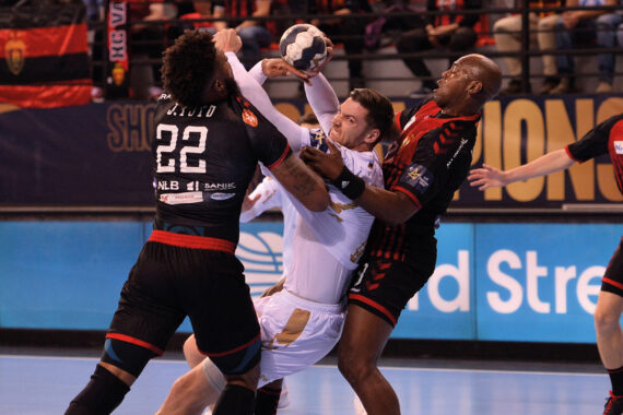 Drei männliche Handballspieler liefern sich einen intensiven Wettkampf; zwei Spieler in schwarz-roten Trikots blockieren einen Spieler in Weiß, der versucht, den Ball zu schießen oder zu passen. Im Hintergrund sind Fahnen und Zuschauer zu sehen.