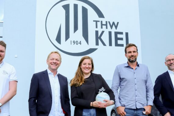 Fünf Personen stehen lächelnd vor einem Gebäude mit einem großen THW Kiel 1904 Logo an der Wand hinter ihnen. Die Person in der Mitte hält eine kleine gläserne Auszeichnung in der Hand.