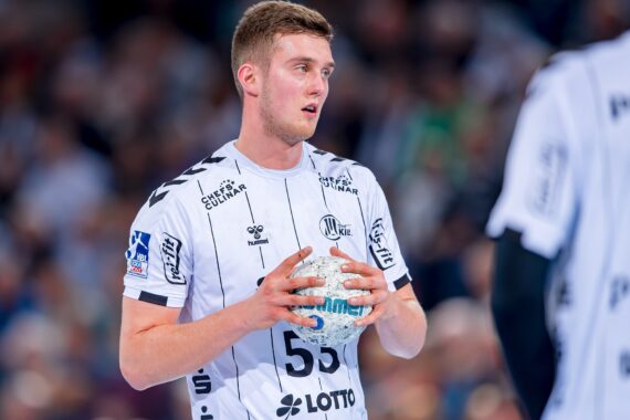 Ein männlicher Handballspieler in einem weißen Trikot hält einen Ball und bereitet sich auf das Spiel vor. Das Publikum ist im Hintergrund verschwommen zu erkennen, und ein anderer Spieler in einem ähnlichen Trikot steht in der Nähe.