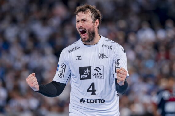 Ein männlicher Handballspieler, der ein weißes Trikot mit der Nummer 4 trägt, jubelt leidenschaftlich mit geballten Fäusten und einem lauten Schrei, während er auf einem Spielfeld steht, mit einer verschwommenen, jubelnden Menge im Hintergrund.