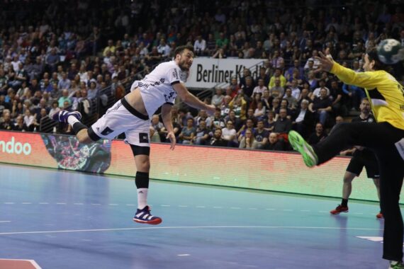 Ein Handballspieler in einem weißen Trikot springt und wirft den Ball in Richtung Tor, während der Torwart in Gelb versucht, ihn zu blocken; eine große Menschenmenge schaut von der Tribüne aus zu.