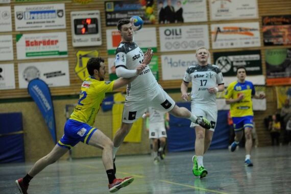Ein Handballspieler in einem weißen Trikot springt mit dem Ball in der Hand in die Luft, während ein Verteidiger in einem gelben Trikot versucht, ihn zu blocken; zwei andere Spieler schauen im Hintergrund in einer Sporthalle zu.