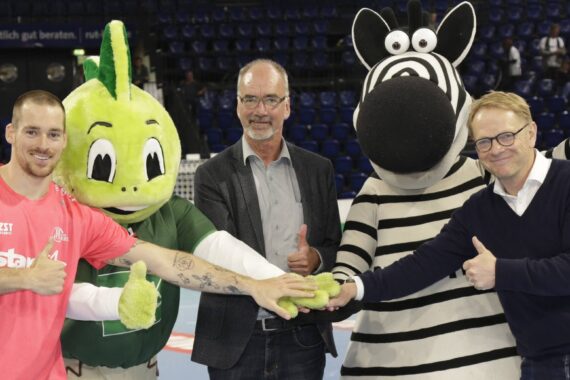Vier Personen, darunter ein Sportmaskottchen in Form eines grünen Drachens und ein weiteres in Form eines Zebras, stehen auf einem Sportplatz, halten die Hände übereinander und zeigen lächelnd die Daumen in Richtung Kamera.