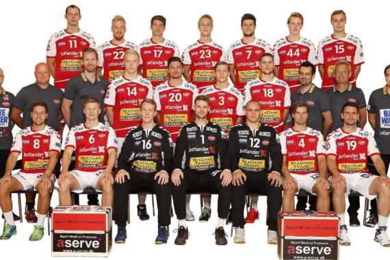 Eine Handballmannschaft von 22 Männern posiert für ein offizielles Gruppenfoto. Sie tragen rote und weiße Trikots, die Trainer und Betreuer sind schwarz gekleidet. Sie sind in drei Reihen vor einem weißen Hintergrund angeordnet, auf dem die Logos der Sponsoren zu sehen sind.