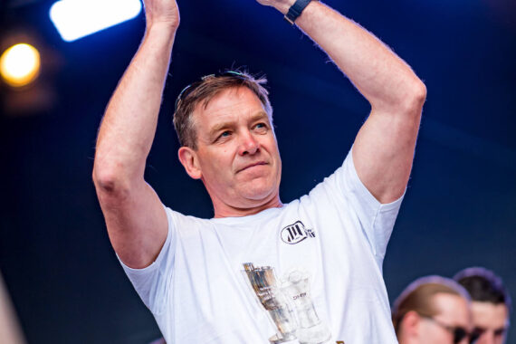 Ein Mann in einem weißen T-Shirt steht auf einer Bühne mit erhobenen Armen und zusammengelegten Händen, als würde er applaudieren. Über ihm leuchtet helles Bühnenlicht, und im Hintergrund sind weitere Personen in ähnlichen Hemden zu sehen.
