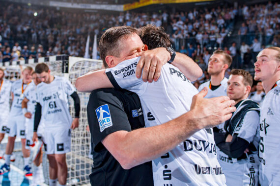 Ein Trainer und ein Spieler umarmen sich freudig auf einem Handballfeld, umgeben von Mannschaftskameraden in weißen Trikots. Die Zuschauer auf der Tribüne schauen zu und feiern einen siegreichen Moment.