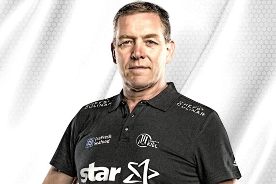 Ein Mann mittleren Alters in einem dunklen Poloshirt mit Sponsorenlogos steht vor einem hellen, gemusterten Hintergrund und schaut selbstbewusst in die Kamera.