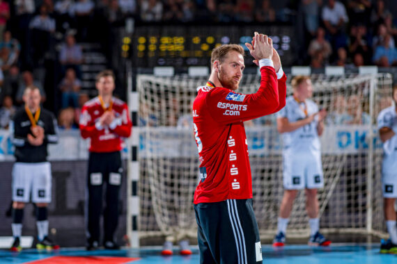 Ein Handballspieler in einem roten Trikot steht vor einem Tor und klatscht, während im Hintergrund andere Spieler in weißen Trikots und eine Menschenmenge zu sehen sind.