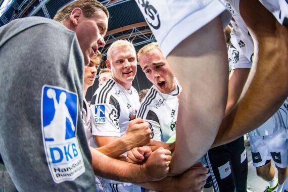Eine Gruppe männlicher Handballspieler in weißen und grauen Uniformen drängt sich zusammen, die Hände in der Mitte gestapelt, um Teamwork und Motivation vor einem Spiel in einer Sporthalle zu zeigen.
