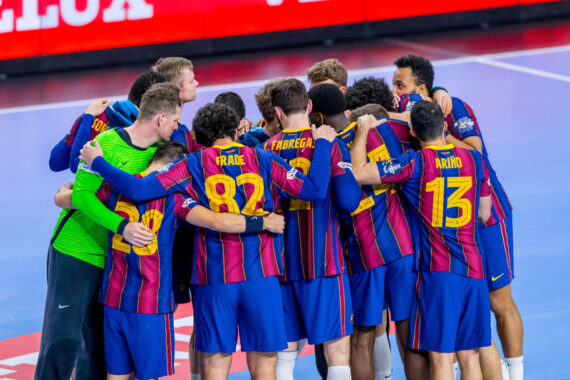 Eine Herren-Handballmannschaft in blau-rot gestreiften Trikots kauert auf einem Hallenplatz zusammen, die Arme umeinander gelegt, und bereitet sich auf ein Spiel vor. Auf dem Rücken der Spieler sind ihre Trikotnummern zu sehen.