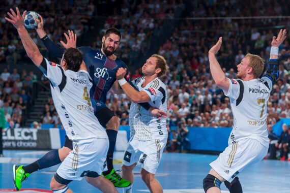 Ein Handballspieler in einem dunklen Trikot springt zum Schuss, während drei Verteidiger in weißen Trikots versuchen, ihn während eines intensiven Spiels in einer überfüllten Halle zu blockieren.