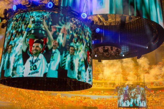 Ein großes Stadion, gefüllt mit jubelnden Fans und hellen Lichtern, zeigt auf großen runden Bildschirmen eine Sportmannschaft, die mit einer Trophäe gefeiert wird. Rauch und leuchtende Farben tragen zur festlichen Atmosphäre bei.