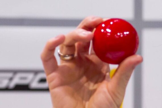 Eine Hand mit einem Ring am Finger hält einen leuchtend roten Ball. Der Hintergrund ist unscharf und enthält weiße und graue Farben und einen Teil des Textes.