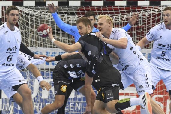 Eine Gruppe männlicher Handballspieler in weißen und schwarzen Trikots kämpft in der Nähe des Tores, während ein Spieler in Schwarz einen Schuss versucht und von einem Spieler in Weiß verteidigt wird. Das Netz und die Spielfeldmarkierungen sind im Hintergrund zu sehen.