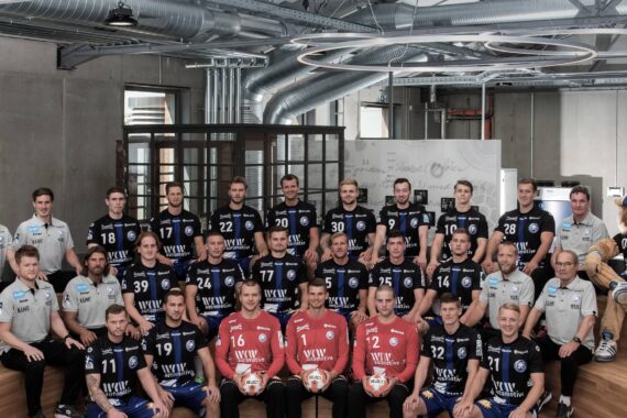 Eine Herrenhandballmannschaft in passenden blauen Trikots posiert in der Halle neben Trainern und Mitarbeitern in hellen Hemden; drei Spieler in roten Trikots halten Handbälle, und ganz rechts sitzt ein blau gekleidetes Maskottchen.
