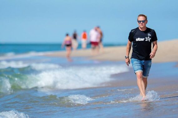 Ein Mann in einem schwarzen T-Shirt und Jeansshorts geht am Strand entlang, die Wellen liegen ihm zu Füßen. Im Hintergrund sind mehrere Menschen zu sehen, die den Strand unter einem strahlend blauen Himmel genießen.