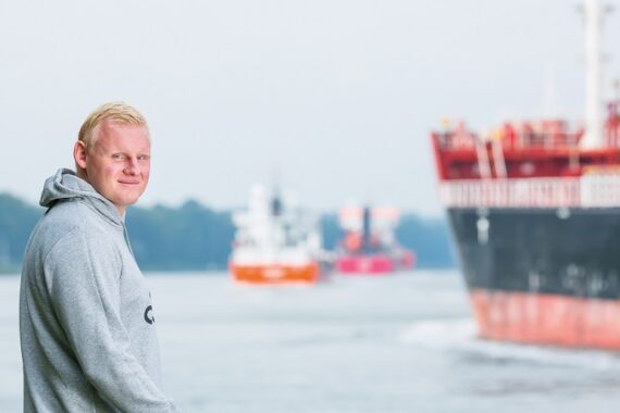 Ein Mann in einem grauen Kapuzenpulli steht am Wasser und schaut in die Kamera, während im Hintergrund an einem bewölkten Tag große Schiffe und Boote vorbeiziehen.