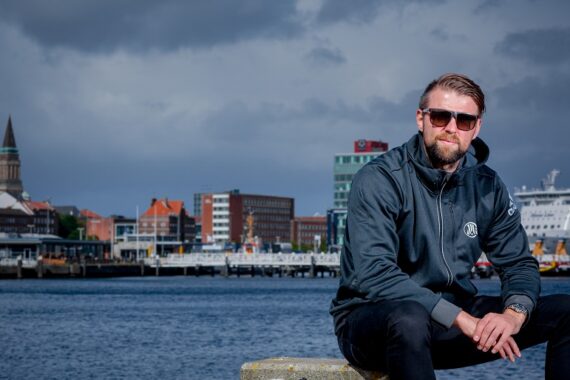 Ein Mann mit Sonnenbrille und dunkler Jacke sitzt am Wasser mit einer Stadtlandschaft, Booten und einem bewölkten Himmel im Hintergrund.