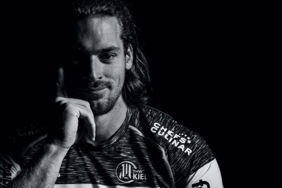 Ein Mann mit langen Haaren und einem leichten Lächeln sitzt in einem dunklen Raum und trägt ein Sporttrikot des THW Kiel. Sein Gesicht ist teilweise beleuchtet, und er stützt seinen Kopf auf seine Hand, wodurch dramatische Schatten entstehen.