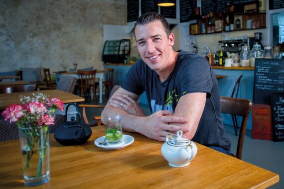 Ein lächelnder Mann sitzt an einem Holztisch in einem gemütlichen Café mit Blumen, einer Teekanne, einem Glas grünem Tee und einem schwarzen Beutel vor sich. Im Hintergrund sind Speisekarten auf Kreidetafeln und Regale mit Flaschen zu sehen.