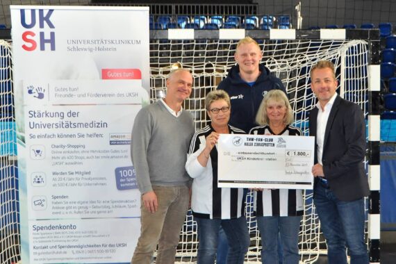Fünf Personen stehen in einer Halle vor einem Fußballnetz und halten einen großen symbolischen Scheck. Auf der linken Seite ist ein Banner des Universitätsklinikums Schleswig-Holstein (UKSH) zu sehen. Alle wirken fröhlich und sind leger gekleidet.